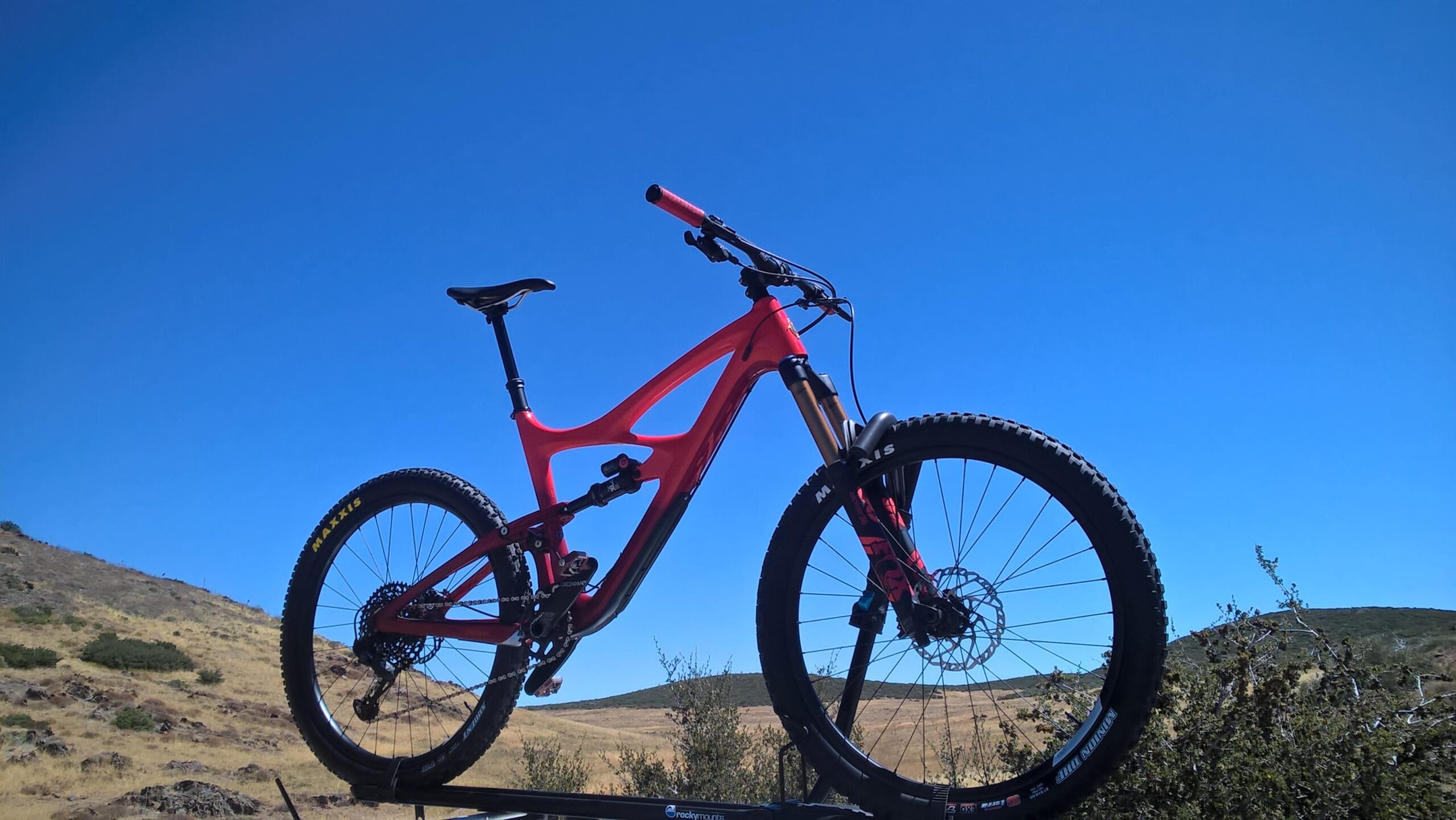 Ibis Mojo HD4: A bright red mountain bike is prominently displayed against a clear blue sky, with rolling hills and sparse vegetation in the background. The bike is positioned on a rack, showcasing its sleek frame and rugged tires designed for outdoor terrain.