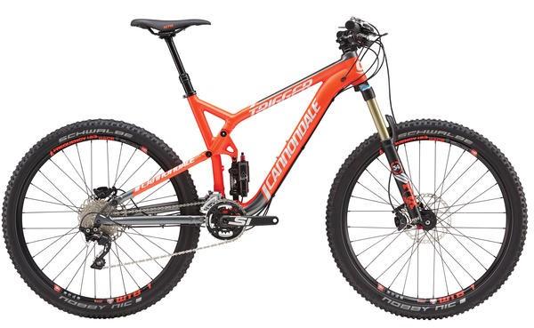 Cannondale Trigger 27.5 3: Mountain bike with a vibrant orange frame, featuring dual suspension and thick, knobby tires designed for rugged terrain. The bike has a sleek, modern design with distinctive branding and high-performance components, including disc brakes and a mix of gears for versatile riding.