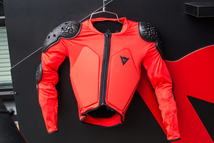 A bright red motorcycle jacket with protective padded shoulders, hanging against a black background. The jacket features a front zipper and a logo on the chest area.