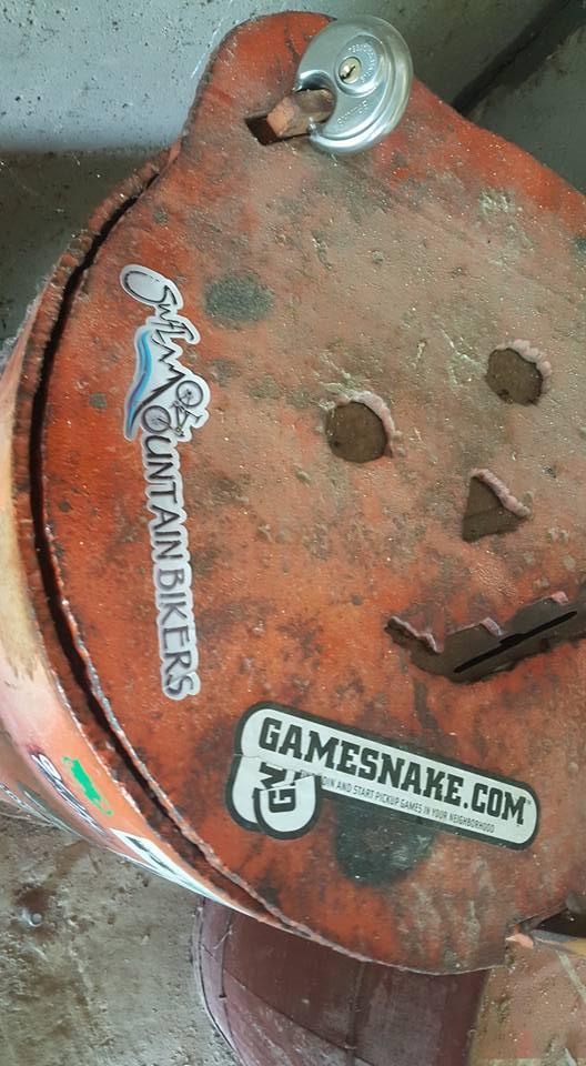 A close-up of a weathered, rusty metal container lid featuring various stickers, including one that reads "Swim Oz Mountain Bikers" and another for "Gamesnake.com." The lid has several holes and a padlock is visible on the upper edge. Dust and wear can be seen on the surface, indicating age and use. Vultures Knob mountain bike trail.