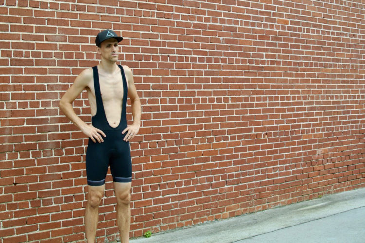 A male athlete stands confidently against a red brick wall, wearing a black cycling bib with a deep cutout and a black cap. He has his hands on his hips, displaying a focused expression, and the background features a smooth concrete surface.
