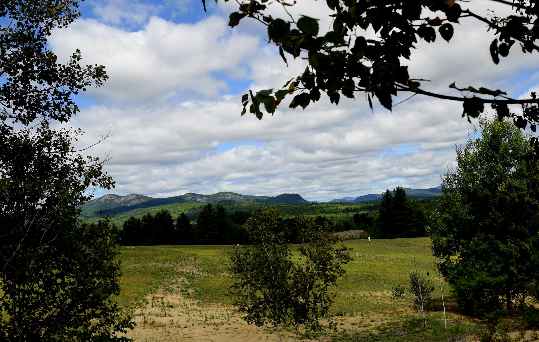 A scenic landscape featuring rolling green hills and distant mountains under a partly cloudy blue sky, framed by trees in the foreground. Ski Bowl Park Bike Trails mountain bike trail.