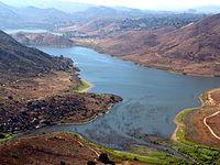 Aerial view of a tranquil lake surrounded by rolling hills and greenery, with a winding shoreline and patches of land visible along the water's edge. Lake Hodges mountain bike trail.