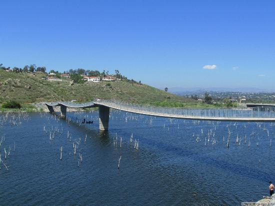 A sleek, modern pedestrian bridge stretches across a calm body of water, flanked by a hillside covered in greenery. The sky is clear with a few scattered clouds, and the water reflects the surroundings. Stumps protrude from the water, hinting at the area's previous vegetation. In the distance, a residential area is visible atop the hill. Lake Hodges mountain bike trail.