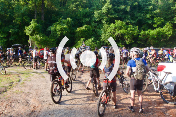 A group of mountain bikers gathered in a wooded area before a ride, with various bicycles parked nearby. Some participants are wearing cycling gear and helmets, while others are socializing and preparing for the event. A natural backdrop of greenery enhances the outdoor atmosphere.