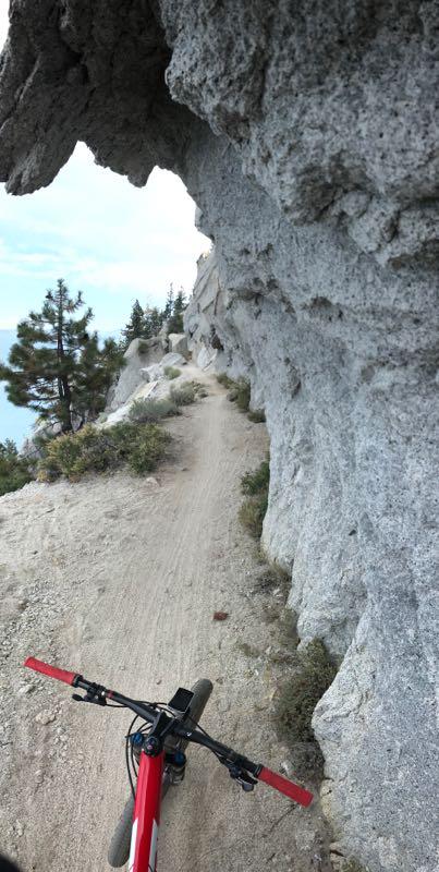 Mountain biking trail viewed from the handlebars of a bike, with a narrow dirt path winding alongside a rocky cliff. Pine trees are visible in the background under a partly cloudy sky. The bike's handlebars and control panel are in the foreground, emphasizing the riding perspective. Flume Trail mountain bike trail.