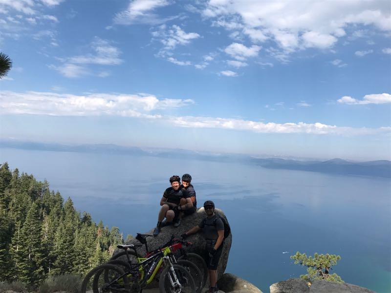Three mountain bikers sitting on a rock with bicycles, overlooking a scenic lake and forested area under a blue sky with clouds. Flume Trail mountain bike trail.