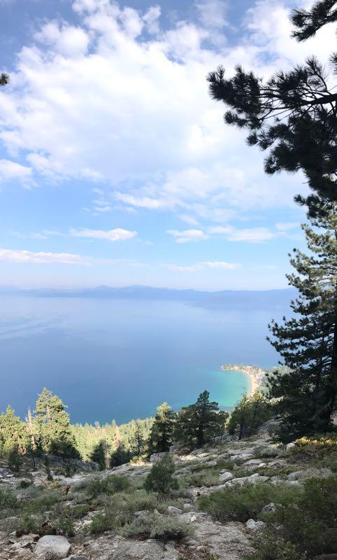 A scenic view of Lake Tahoe, showcasing the expansive blue water under a partly cloudy sky, framed by lush green trees and rocky terrain. The landscape includes a glimpse of the shoreline and distant mountains in the background. Flume Trail mountain bike trail.