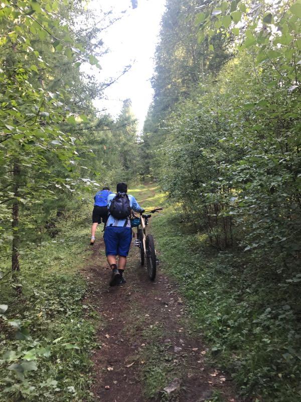 Two individuals walking along a narrow dirt path through a lush green forest, one carrying a bicycle. The terrain is surrounded by dense trees and underbrush, suggesting a nature trail used for hiking or biking. Bright natural light filters through the foliage above. Zaisan Chutes mountain bike trail.
