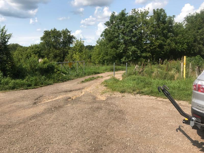 A gravel road leading to a gate with a warning sign, surrounded by lush greenery and trees under a partly cloudy sky. A vehicle is parked nearby, with the rear tow hitch visible. The scene depicts a rural setting with a clear division between the road and the surrounding vegetation. FDR Park mountain bike trail.