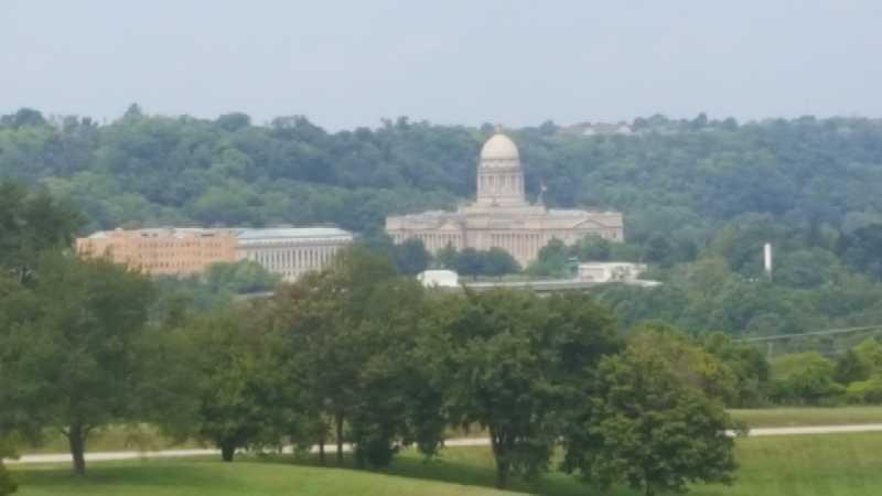 A view of a large building with a prominent dome set against a green hillside, surrounded by trees and clear blue skies. The image captures the architectural details of the structure and the natural landscape in the foreground. Capital View mountain bike trail.