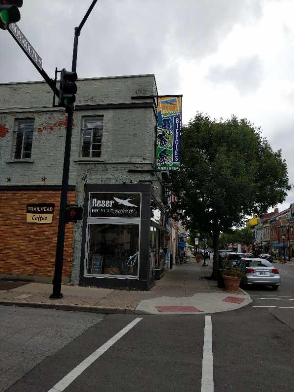 A street view of a small town featuring a storefront labeled "Reser Bicycles." The building has a light-colored exterior with brick accents, and a colorful banner hanging above the entrance. A tree is visible in front of the shop, and a coffee shop named "Trailhead Coffee" is nearby. The image captures a cloudy sky and parked cars along the street.