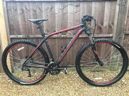 Specialized Rockhopper Comp Disc: A black and red mountain bike leaning against a wooden fence, featuring thick tires, front suspension, and a modern frame design. The bike is positioned on a gravel surface.