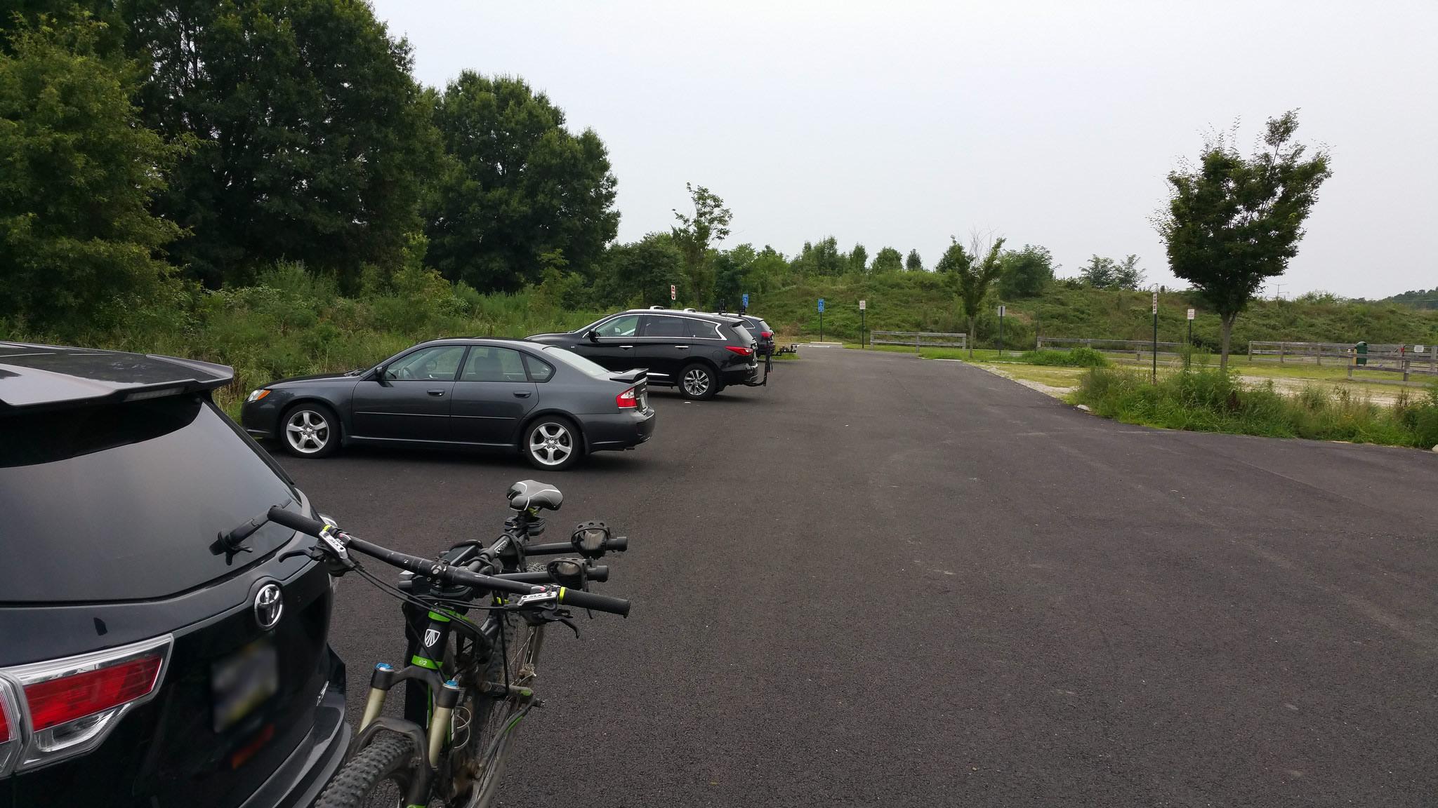 A parking area with several vehicles, including a car parked in the foreground with a bike attached to its rack. The background features greenery and trees, with a few parking signs and an open space. The atmosphere is overcast, suggesting a cloudy day. Laurel Hill Park mountain bike trail.