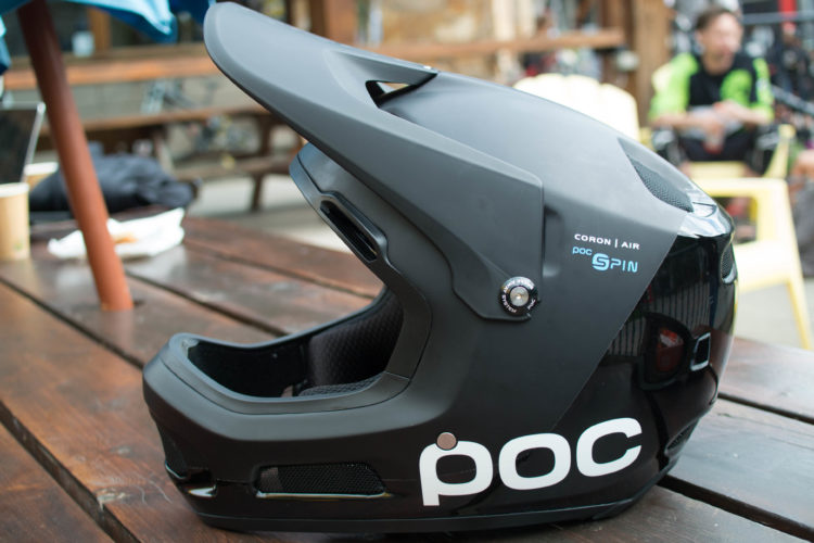 A black mountain biking helmet with a sleek design, featuring ventilation ports and the logo "poc" prominently displayed. The helmet is positioned on a wooden table, with a blurred background showing outdoor furniture and other cyclists.