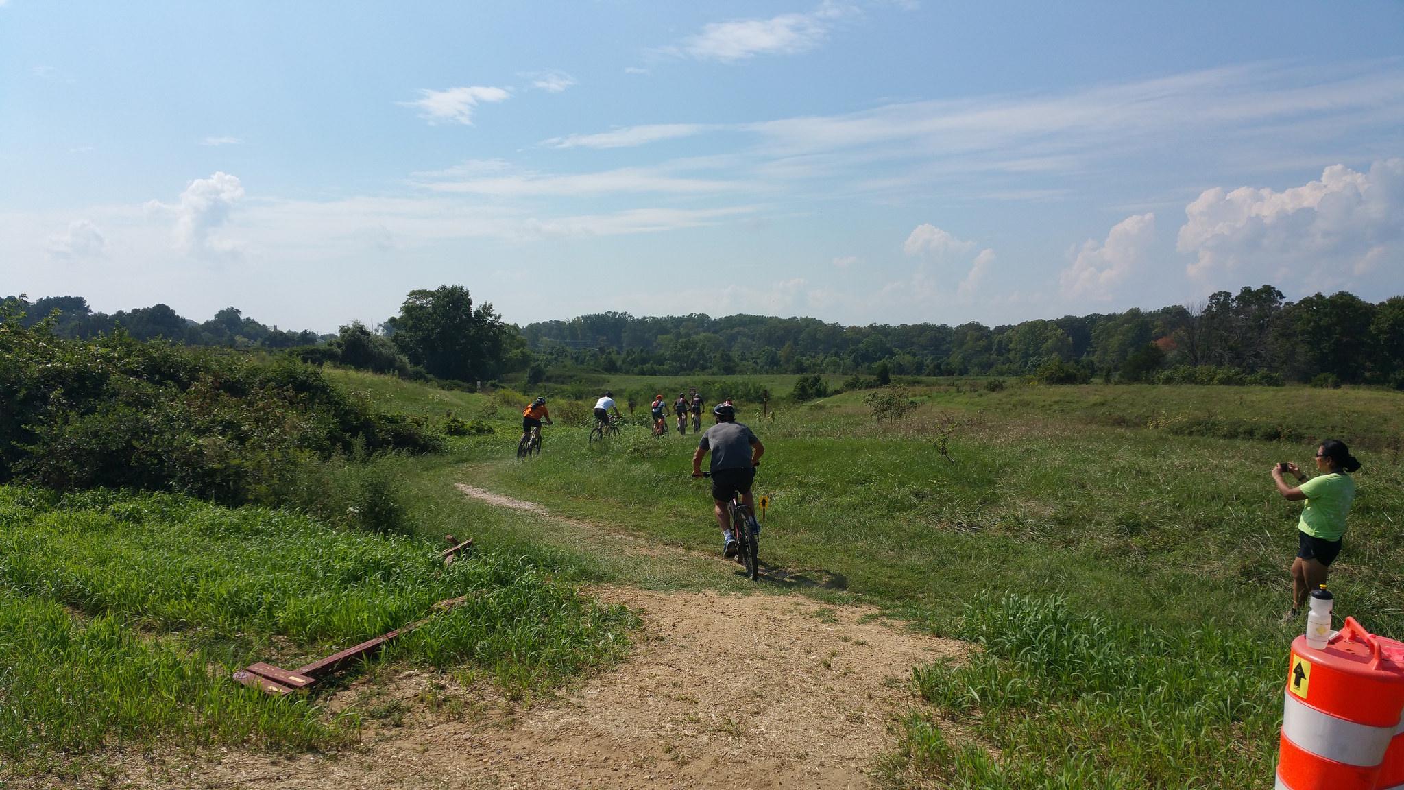 A group of cyclists riding on a dirt trail through a lush green landscape under a clear blue sky. In the foreground, a person in a green shirt is taking a photo, while others ride along the winding path in the background. Laurel Hill Park mountain bike trail.