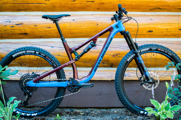 A mountain bike with a maroon and light blue frame, featuring thick tires with aggressive tread patterns, is positioned against a rustic log wall. Green foliage is visible in the foreground, and the bike showcases modern components, including a dropper seat post and a rear shock.