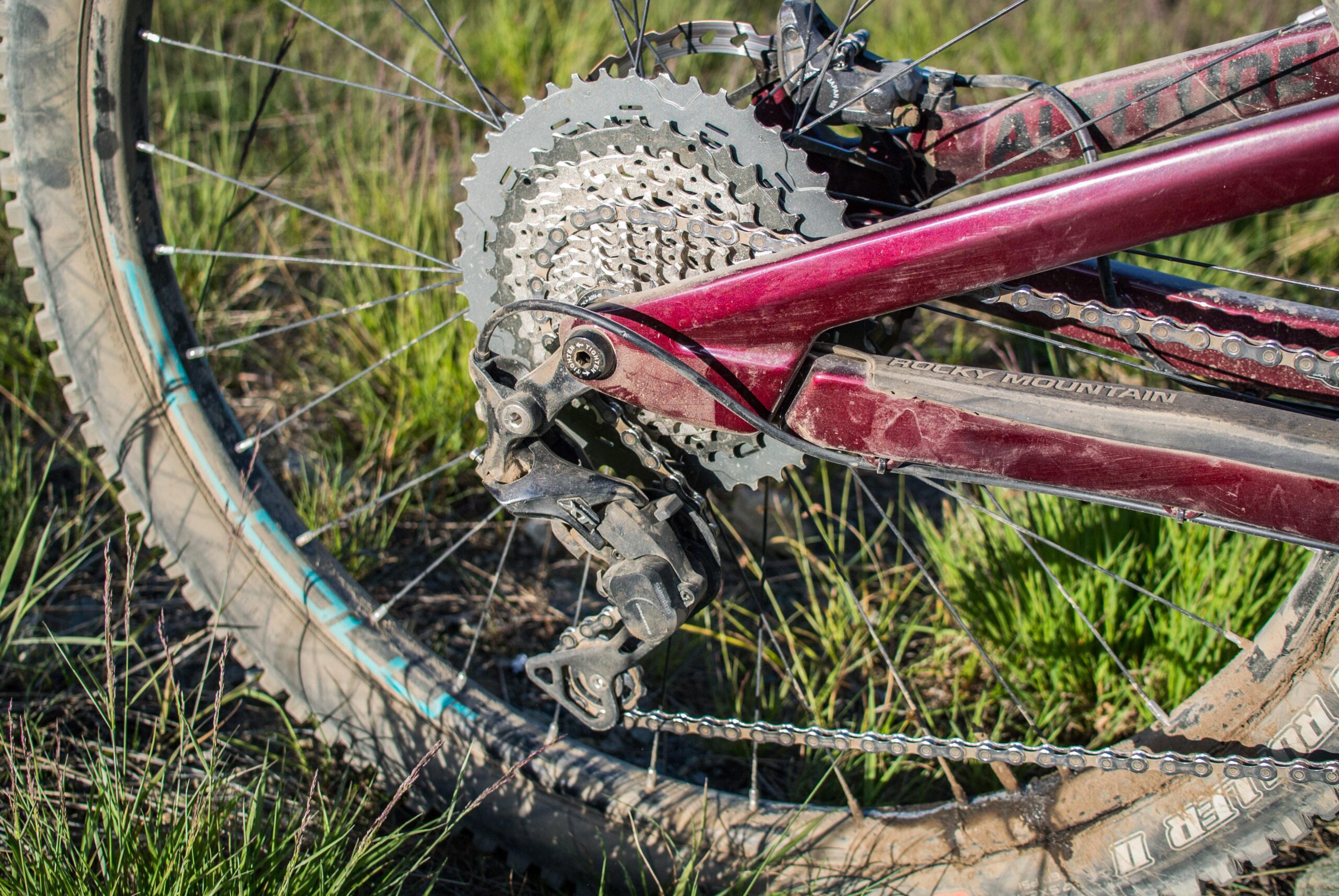 Rocky Mountain Altitude: Close-up view of a mountain bike's rear derailleur and gears, showcasing the chain and cassette against a background of green grass. The bike's frame is maroon, and the components appear slightly dusty, indicating recent use on trails.