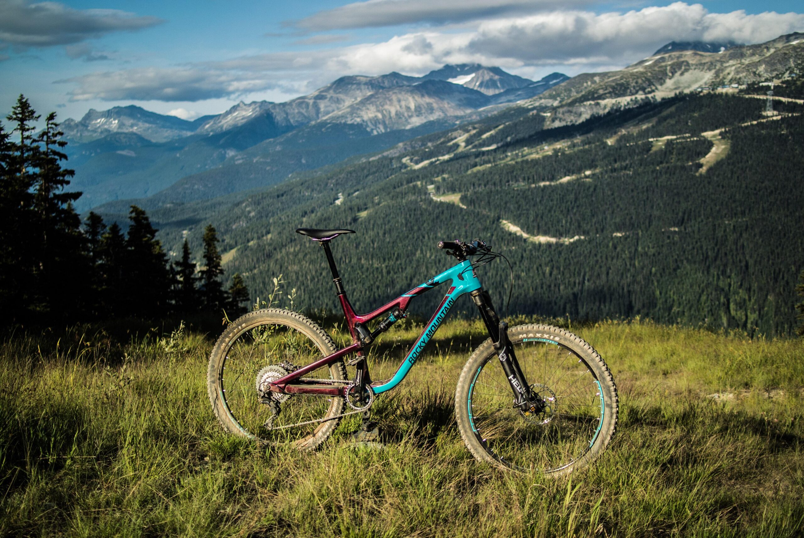 Rocky Mountain Altitude: Mountain bike resting on grass with a backdrop of mountains and a blue sky. The bike features a color scheme of deep red and turquoise, and the surrounding landscape includes dense forests and rolling hills.
