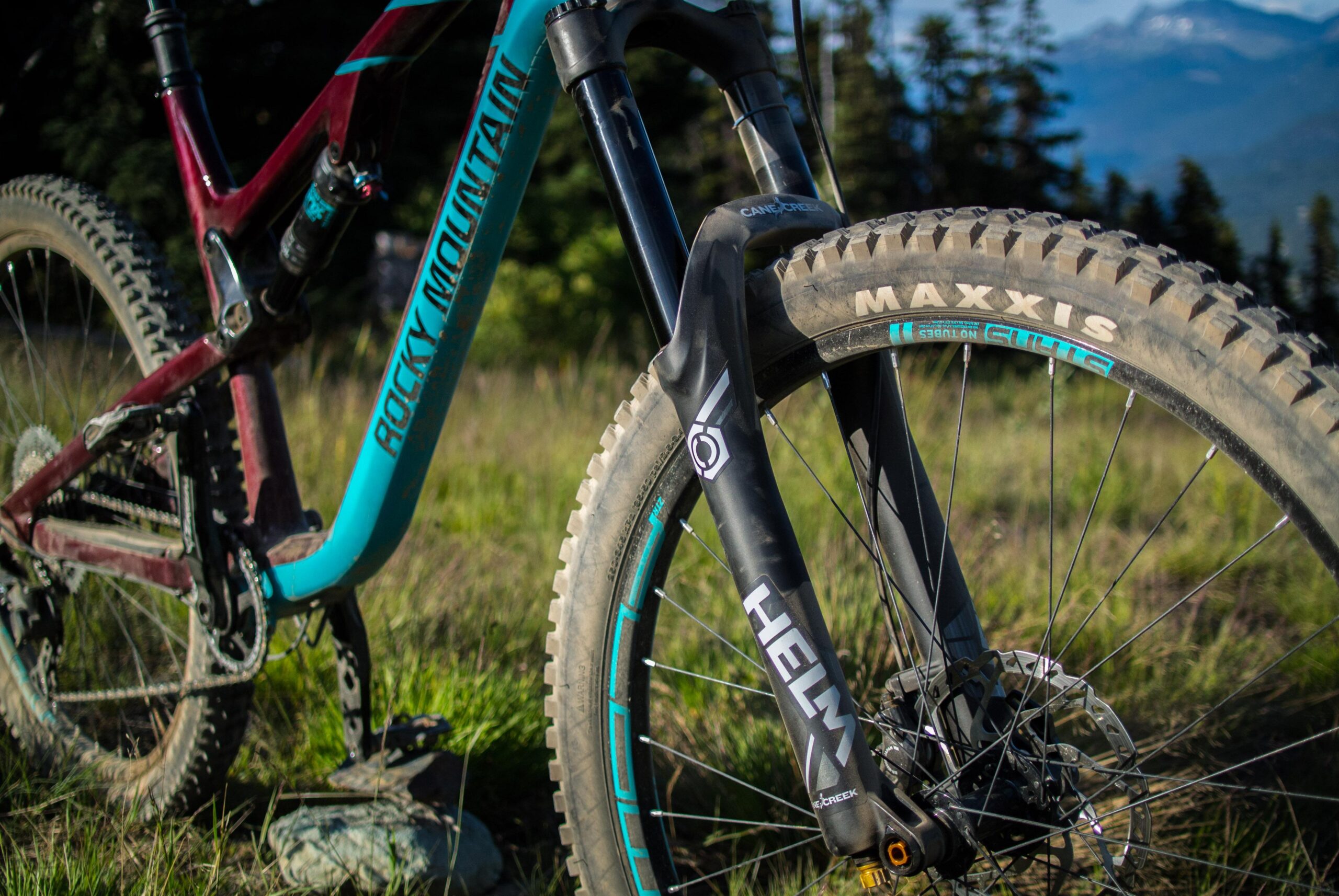 Rocky Mountain Altitude: Close-up of a mountain bike featuring a Rocky Mountain frame and Helm suspension fork, with a Maxxis tire in a natural outdoor setting. The bike is partially in focus against a grassy background with trees and mountains visible in the distance.