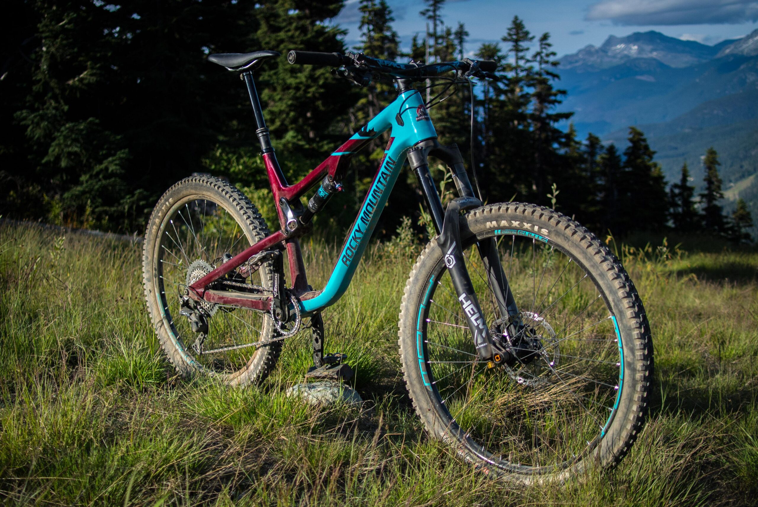 Rocky Mountain Altitude: A mountain bike with a turquoise and maroon frame parked on grassy terrain, surrounded by trees and mountains in the background. The bike features a black front fork and thick tires designed for off-road riding.