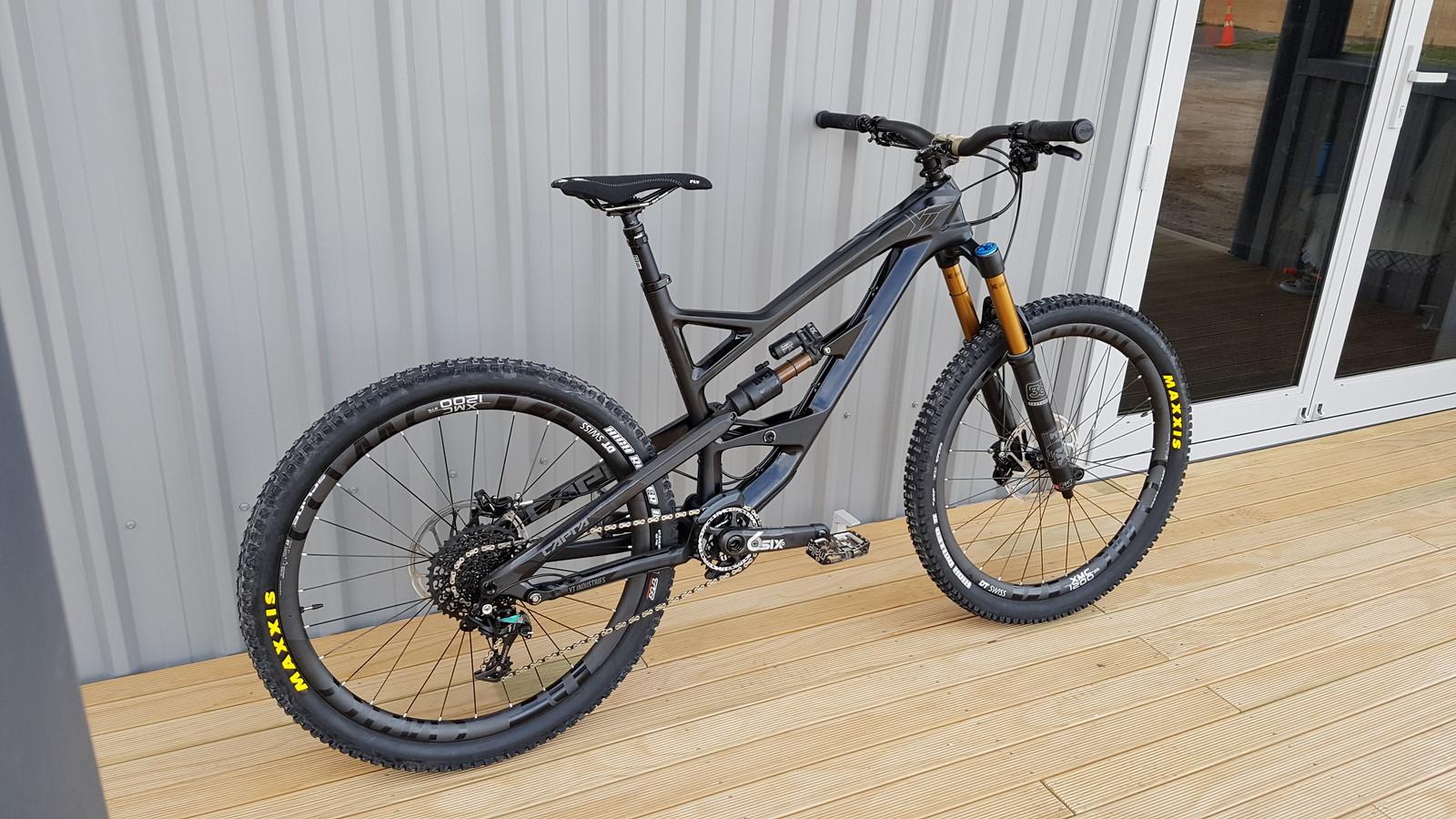 YT Industries Capra Carbon: A sleek black mountain bike with thick tires and a dual suspension system, positioned on a wooden deck in front of a metal wall. The bike features prominent branding on the wheels and a high-end fork with gold components, suggesting it's built for performance on rough terrain. The background includes a glass door, indicating an indoor setting.