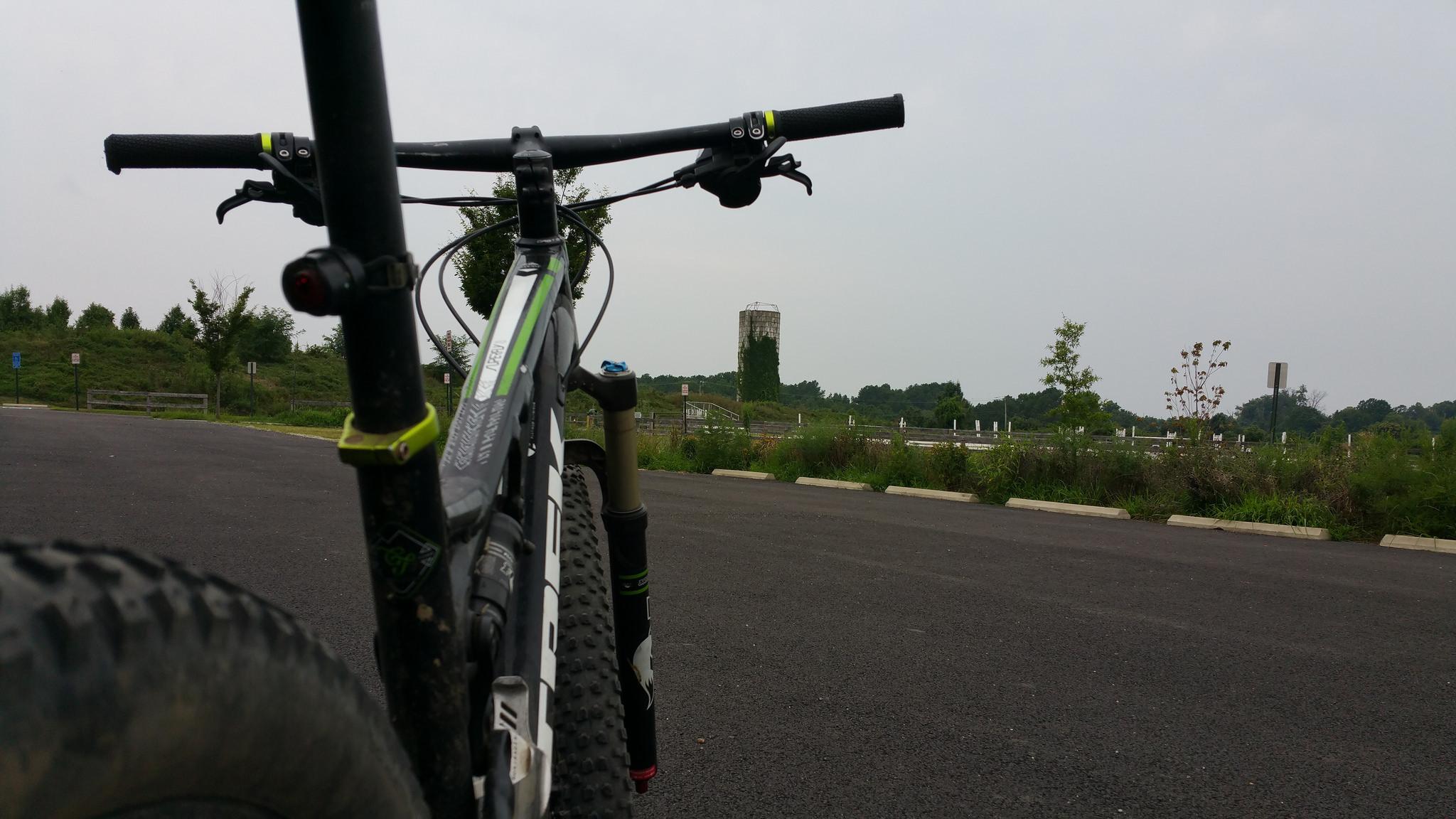 A close-up view of a mountain bike positioned on a paved surface, with a backdrop of a grassy area and a distant silo. The image captures the bike's handlebars and front tire, emphasizing the rugged design. Overcast skies are visible above, indicating a cloudy day. Laurel Hill Park mountain bike trail.