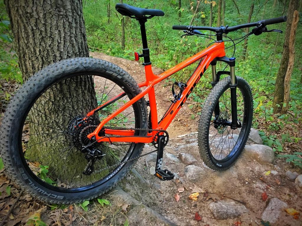 A vibrant orange mountain bike leaned against a tree on a woodland trail, showcasing its sturdy tires and detailed components. The background features lush greenery and a winding path made of dirt and rocks, indicating an adventurous outdoor setting.