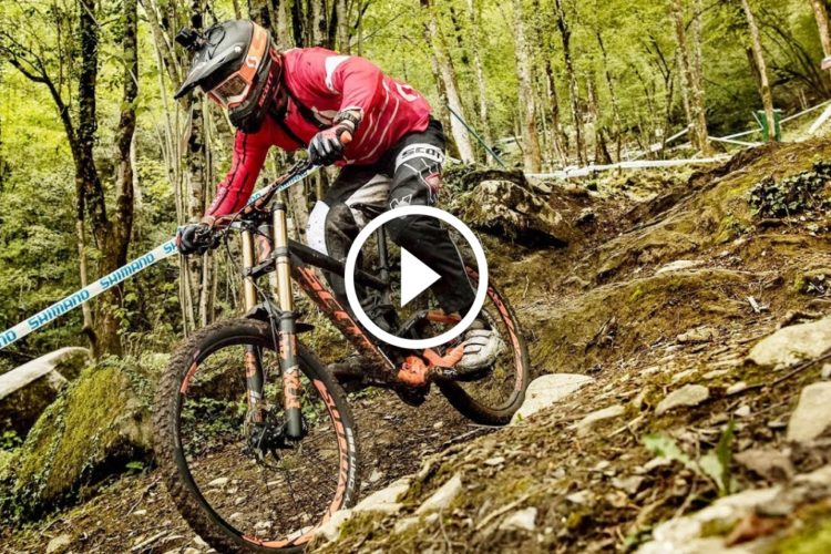A mountain biker navigating a rocky trail in a forested area, wearing a red jersey and helmet while riding a black and orange bike. The scene captures the dynamic motion and rugged terrain of the sport.
