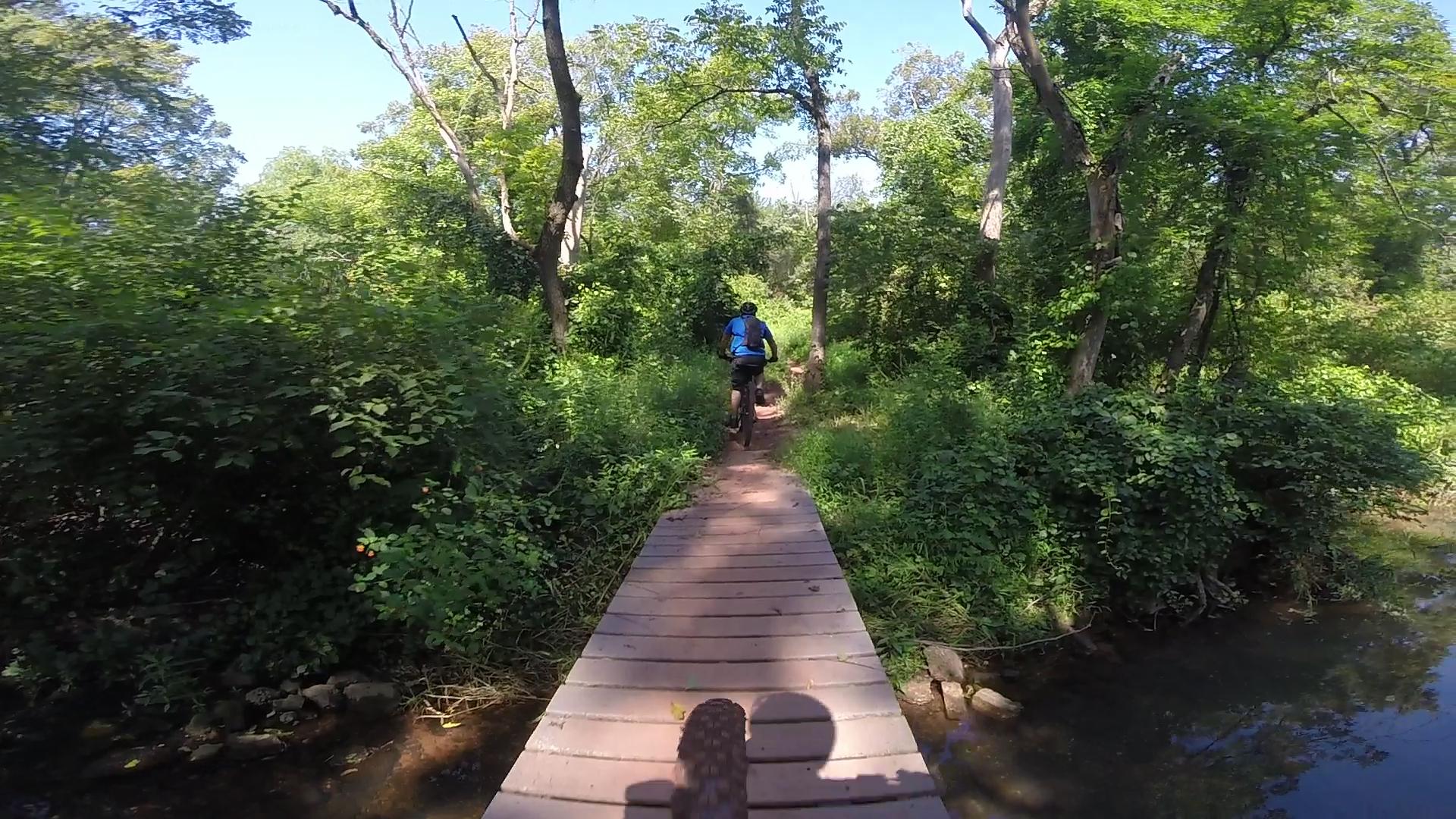 A cyclist rides over a wooden bridge surrounded by lush greenery, with trees and bushes lining the path. The bright blue sky is visible above, indicating a sunny day. Six Mile Run mountain bike trail.