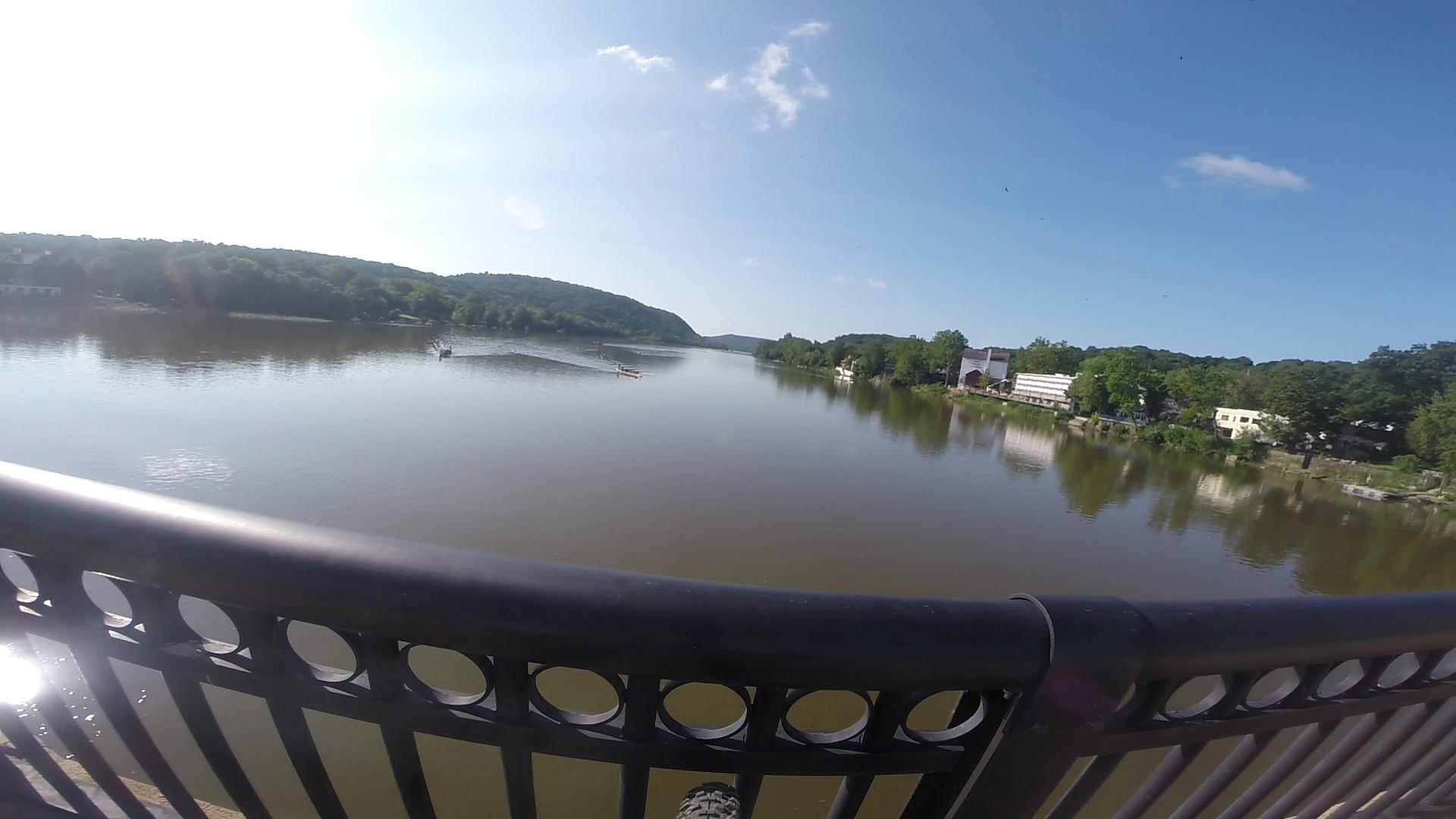 A scenic view from a bridge overlooking a calm river surrounded by lush green hills and trees, with some buildings visible along the shore under a clear blue sky. Bridge To Bridge - D&r Canal mountain bike trail.