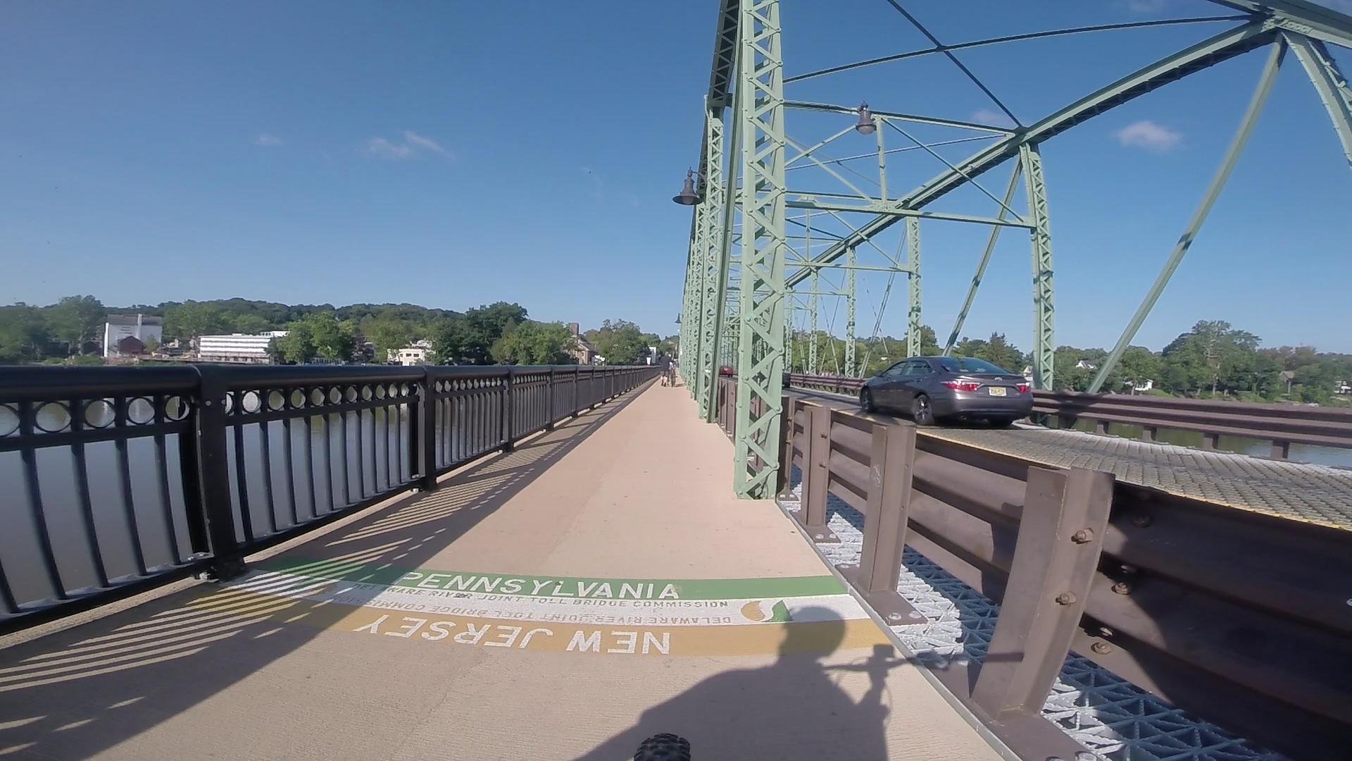 A view of a bridge with green metal supports, featuring a pedestrian walkway marked with “PENNSYLVANIA” and “NEW JERSEY” signage. A car is driving on the adjacent lane, with a river visible to the side and trees and buildings in the background under a clear blue sky. Bridge To Bridge - D&r Canal mountain bike trail.