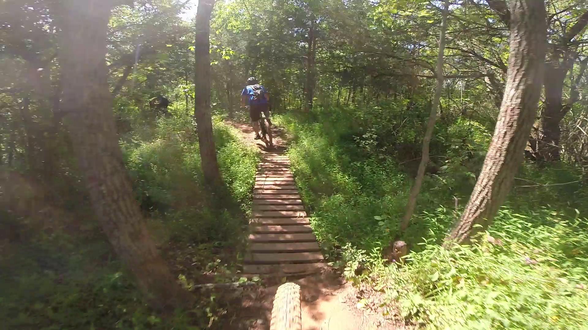 A mountain biker riding along a wooden plank trail in a dense forest, surrounded by trees and greenery, with sunlight filtering through the leaves. Six Mile Run mountain bike trail.