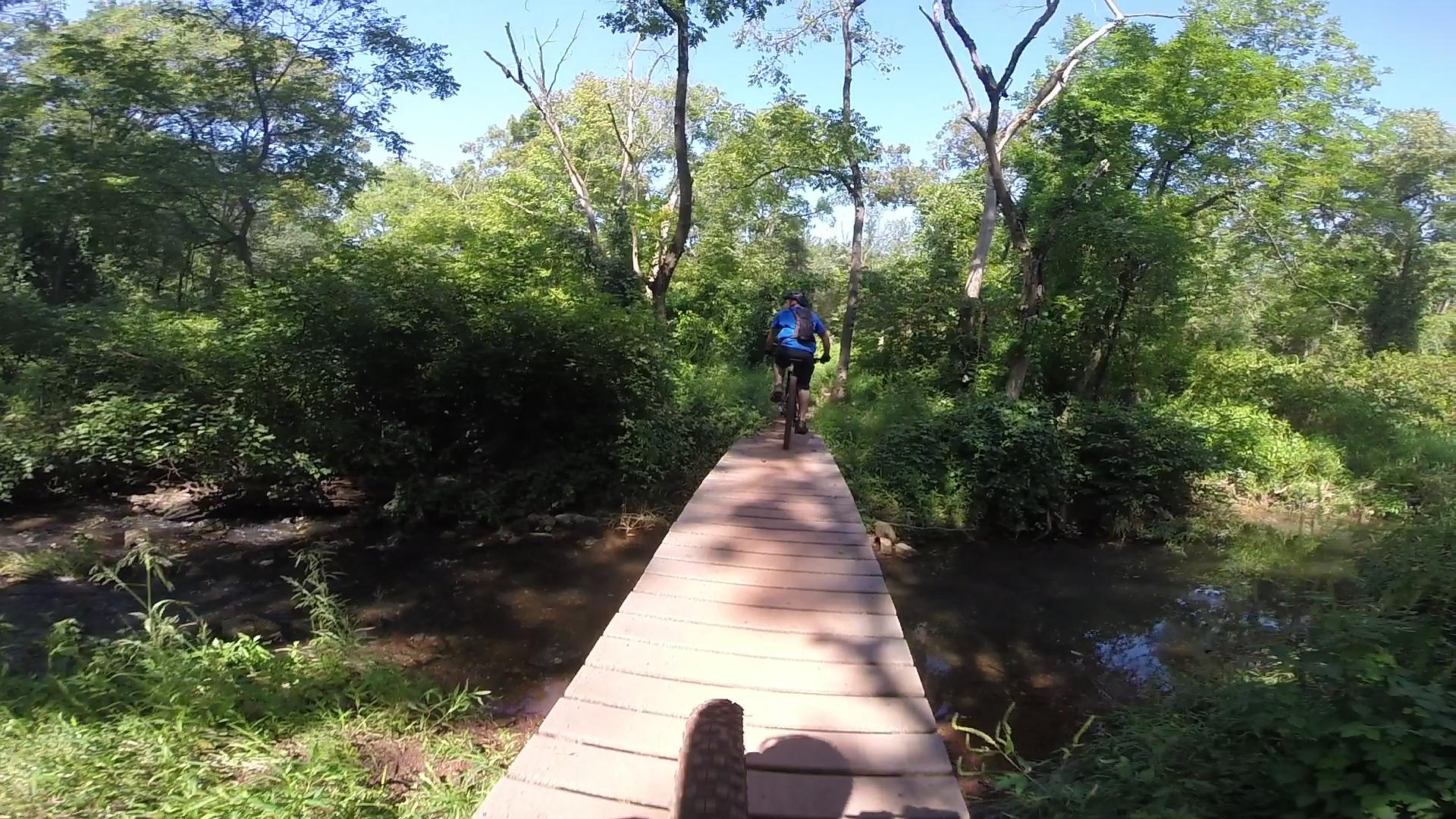 A person riding a mountain bike across a wooden bridge in a lush green forest. Sunlight filters through the trees, illuminating the pathway that leads over a small stream. Six Mile Run mountain bike trail.