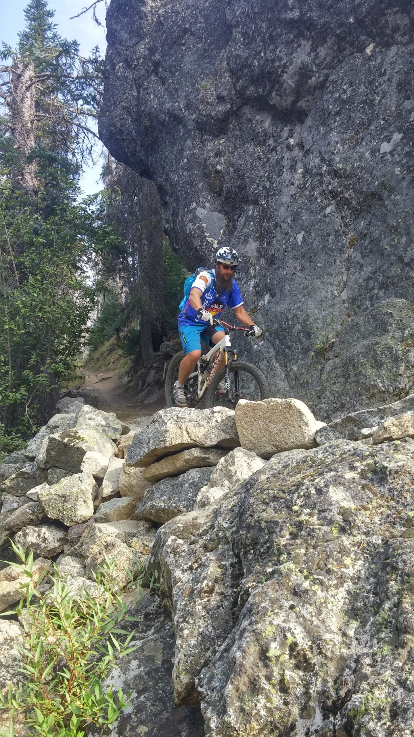 A mountain biker navigating a rocky trail in a forested area, with large boulders and trees in the background. Loon Lake Trail mountain bike trail.