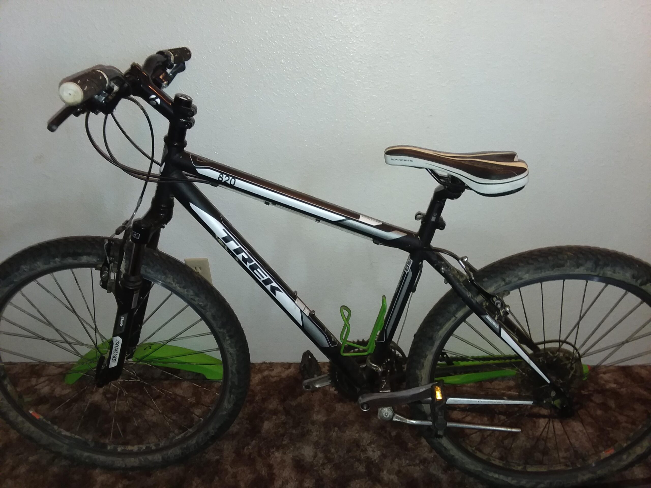 Trek 820: A black and white Trek mountain bike with green accents, featuring a textured seat and handlebars equipped with grips and a mini light. The bike is parked on a carpeted surface, and its wheels show signs of regular use.