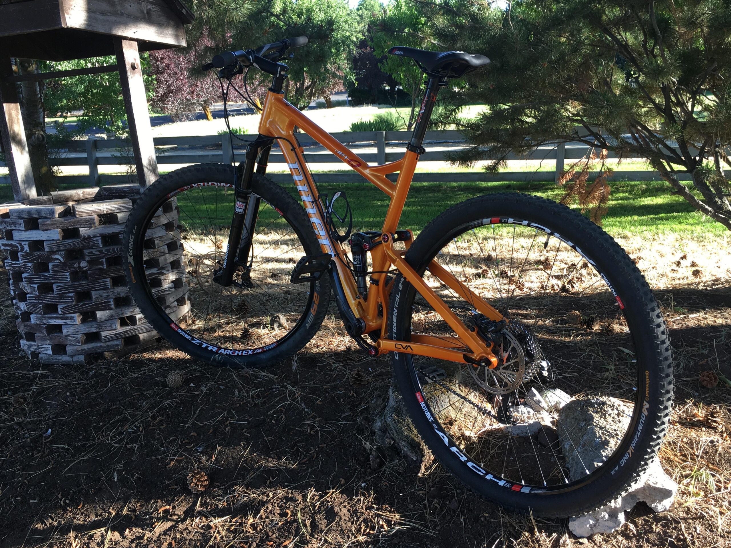 Niner Jet 9: A bright orange mountain bike parked next to a wooden well, set against a backdrop of green grass and trees. The bicycle features thick tires with visible brand labels and a sturdy frame design, showcasing its off-road capability. Sunlight filters through the trees, casting dappled shadows on the ground.