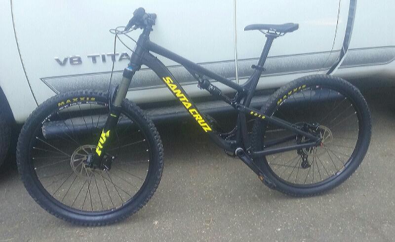 Santa Cruz 5010: A black mountain bike with yellow accents, featuring the brand name "Santa Cruz" on the frame. The bike has wide tires and a sturdy design, positioned next to a white pickup truck.