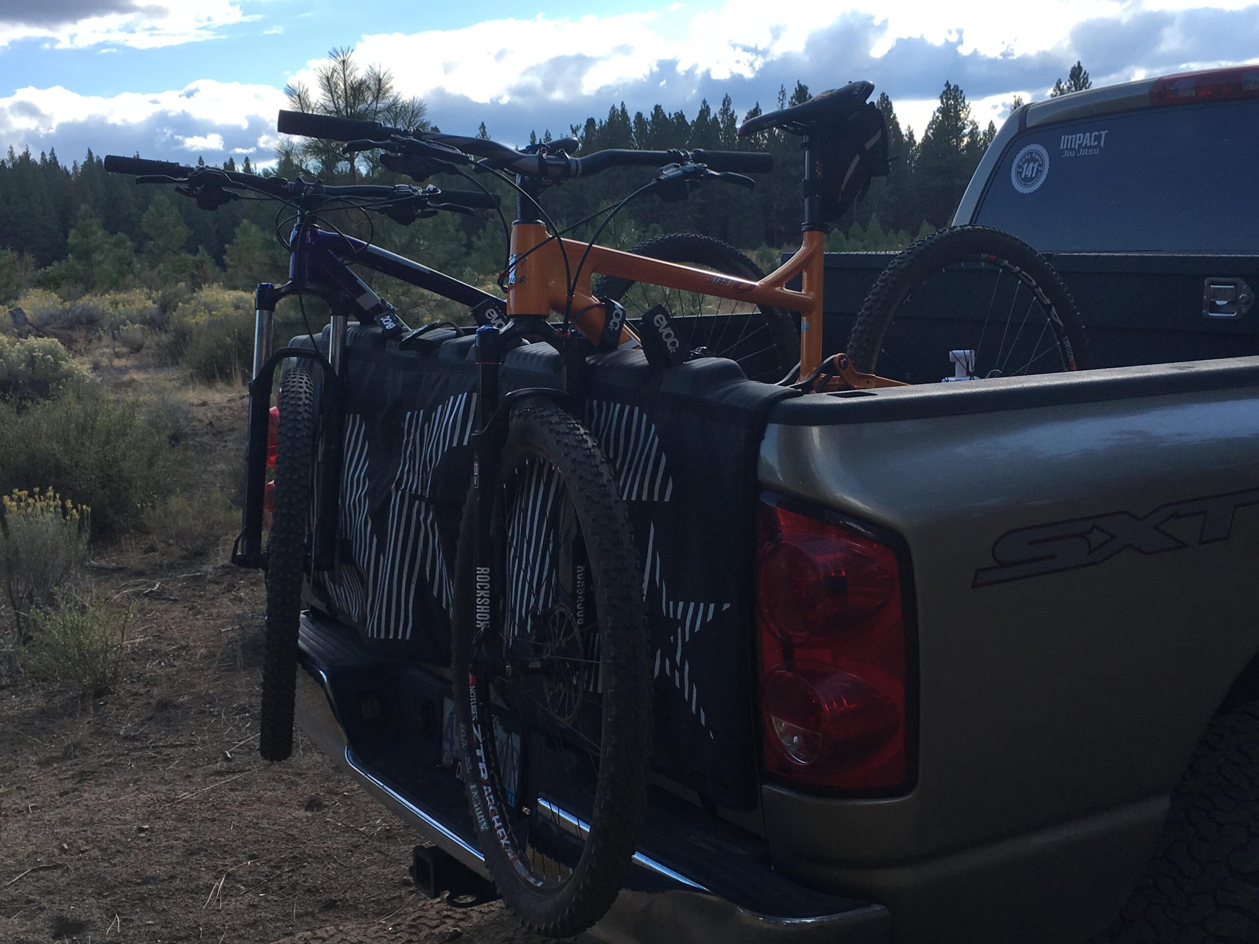 Niner Jet 9: A close-up view of the back of a pickup truck, showcasing two mountain bikes secured to a bike rack in the truck bed. The bikes are positioned upright, one in purple and one in orange, against a backdrop of trees and a cloudy sky. The truck features a silver exterior with visible taillights and a sticker on the back window. The ground is natural terrain, suggesting an outdoor setting.