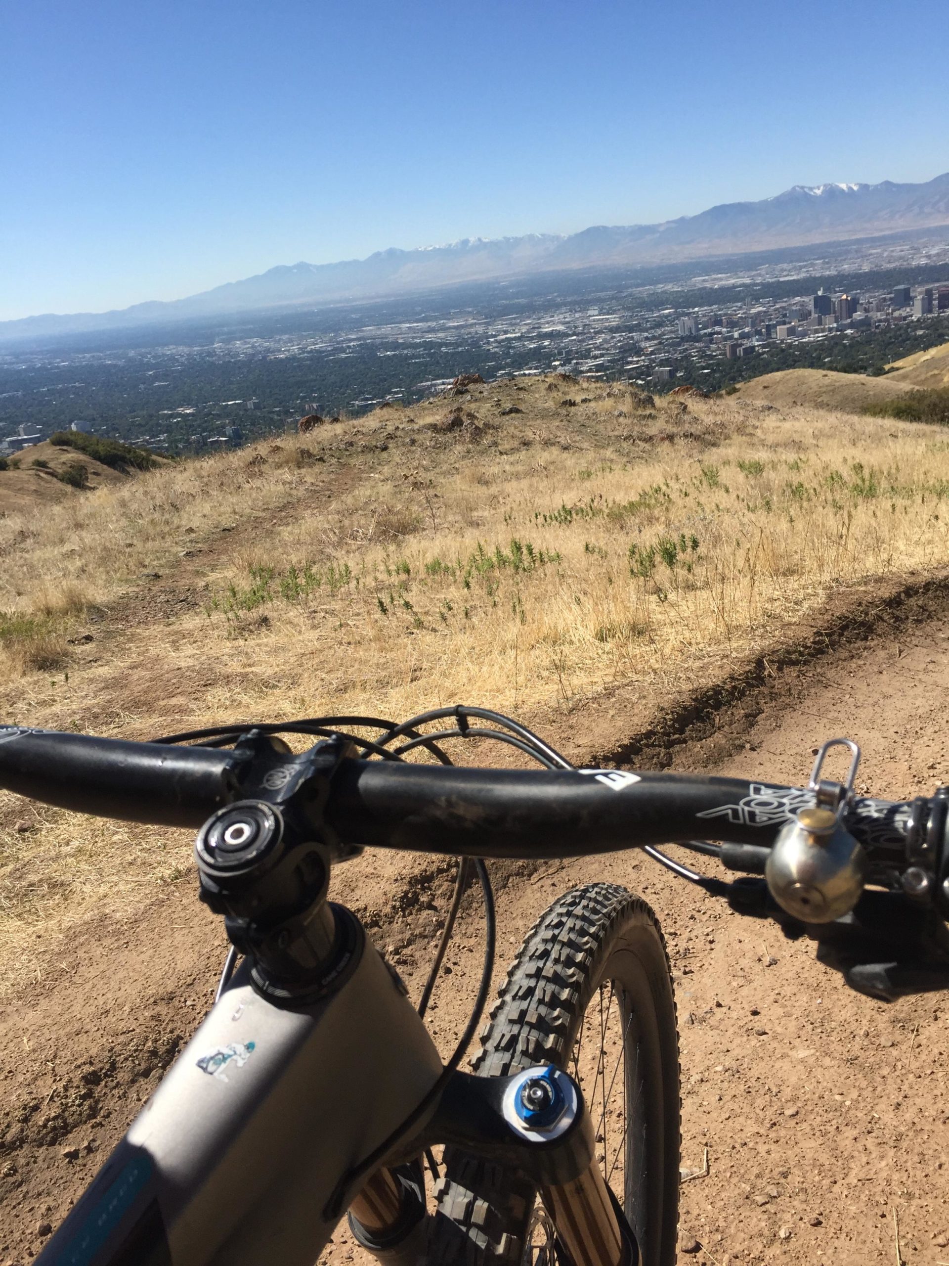 A view from the handlebars of a mountain bike, overlooking a city with a backdrop of mountains under a clear blue sky. The foreground shows the bike’s controls and tire on a dirt trail, while the scenery features a blend of urban and natural landscapes. Mt. Wire mountain bike trail.