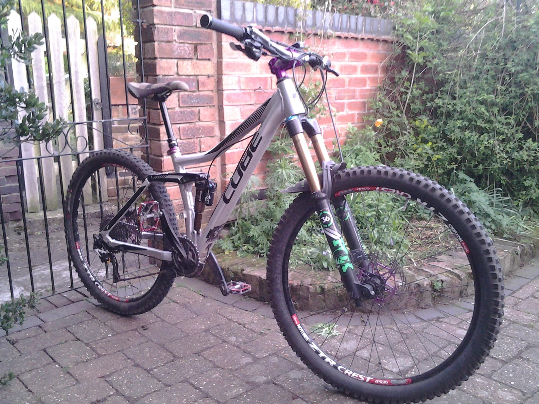 Cube Cube Stereo 160 HPA SL: A silver and black Cube mountain bike is positioned on a brick pathway. The bike features thick, knobby tires and a dual-suspension frame, with a prominent front fork. In the background, a metal gate and brick wall are visible, alongside some greenery and flowers.