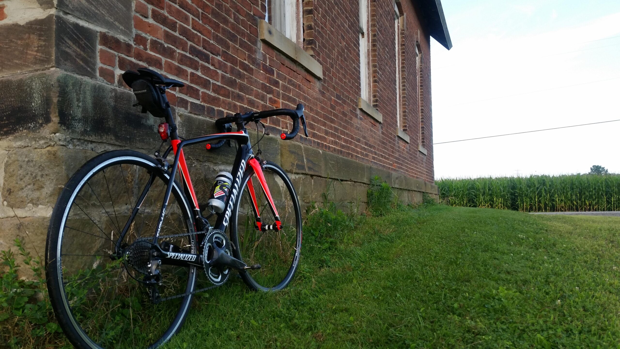 A black and red road bike with a water bottle attached, leaning against a brick wall beside a grassy area, with cornfields visible in the background under a clear sky.