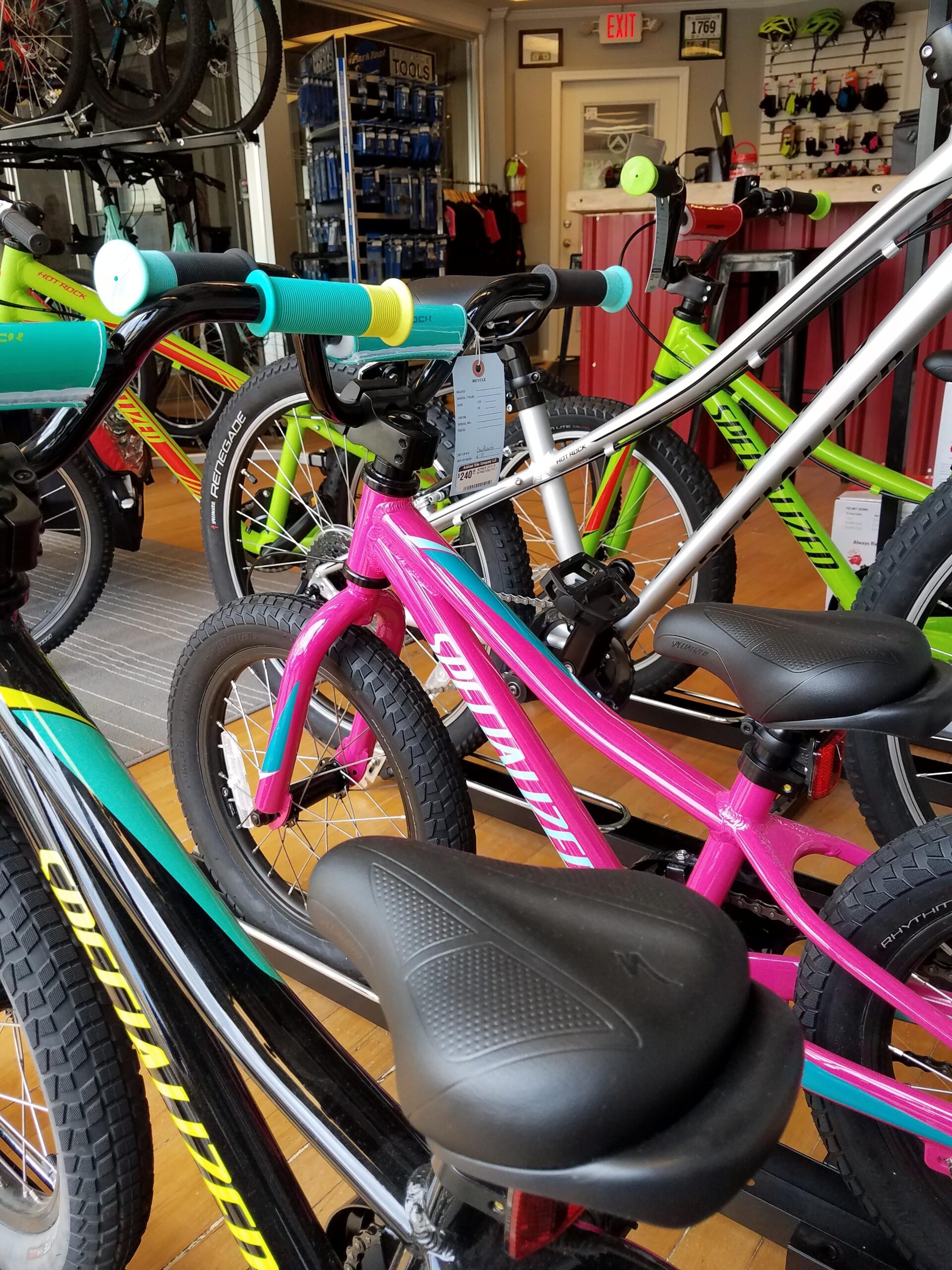 A colorful display of bicycles in a shop, featuring various models and sizes. In the foreground, a pink bicycle stands out with distinctive handlebars and seat, while other bikes in shades of green and silver are visible in the background. The shop interior includes a wall of bike accessories and tools.