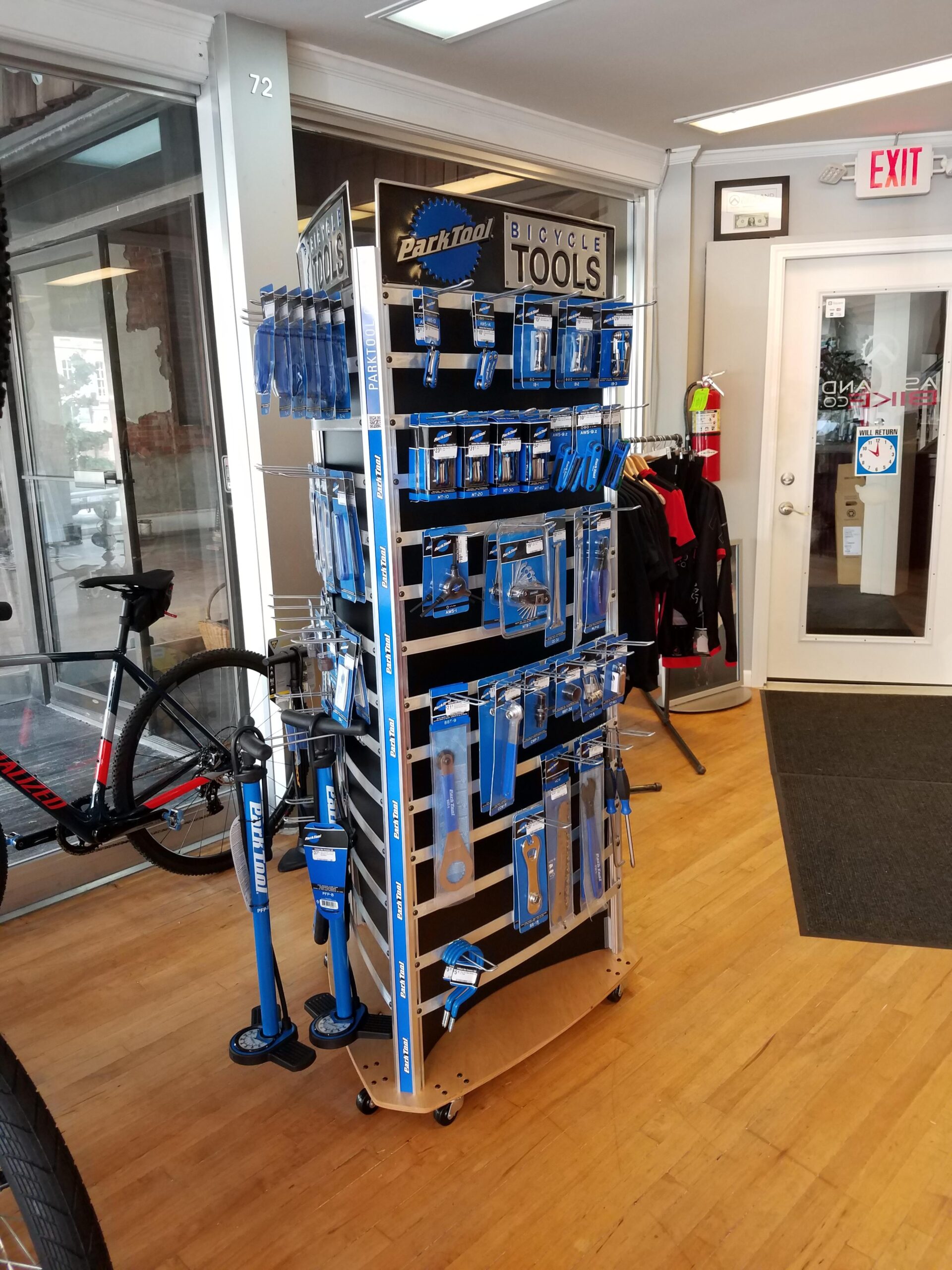 A display rack showcasing various bicycle tools from Park Tool, featuring items such as tire pumps, wrenches, and repair tools, inside a bicycle shop. In the background, there is a bicycle leaning against the wall and an exit door visible. The shop has a wooden floor and a bright, welcoming atmosphere.
