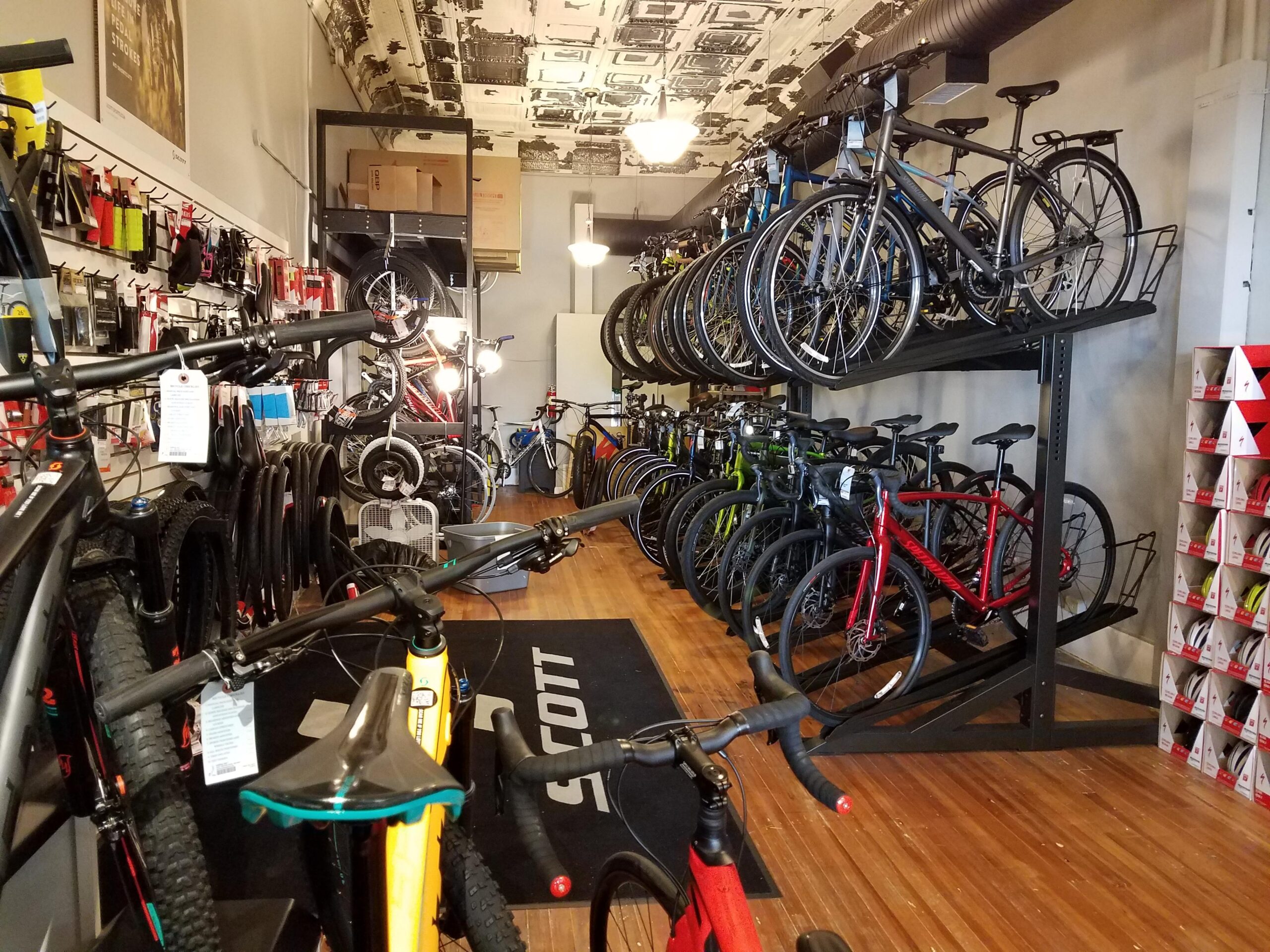 A bicycle shop interior featuring a variety of bicycles on display, including mountain bikes and road bikes. Display racks hold bikes in various colors, and accessories are visible on the walls. The shop has hardwood flooring and a stylish ceiling, creating a vibrant atmosphere for cycling enthusiasts.