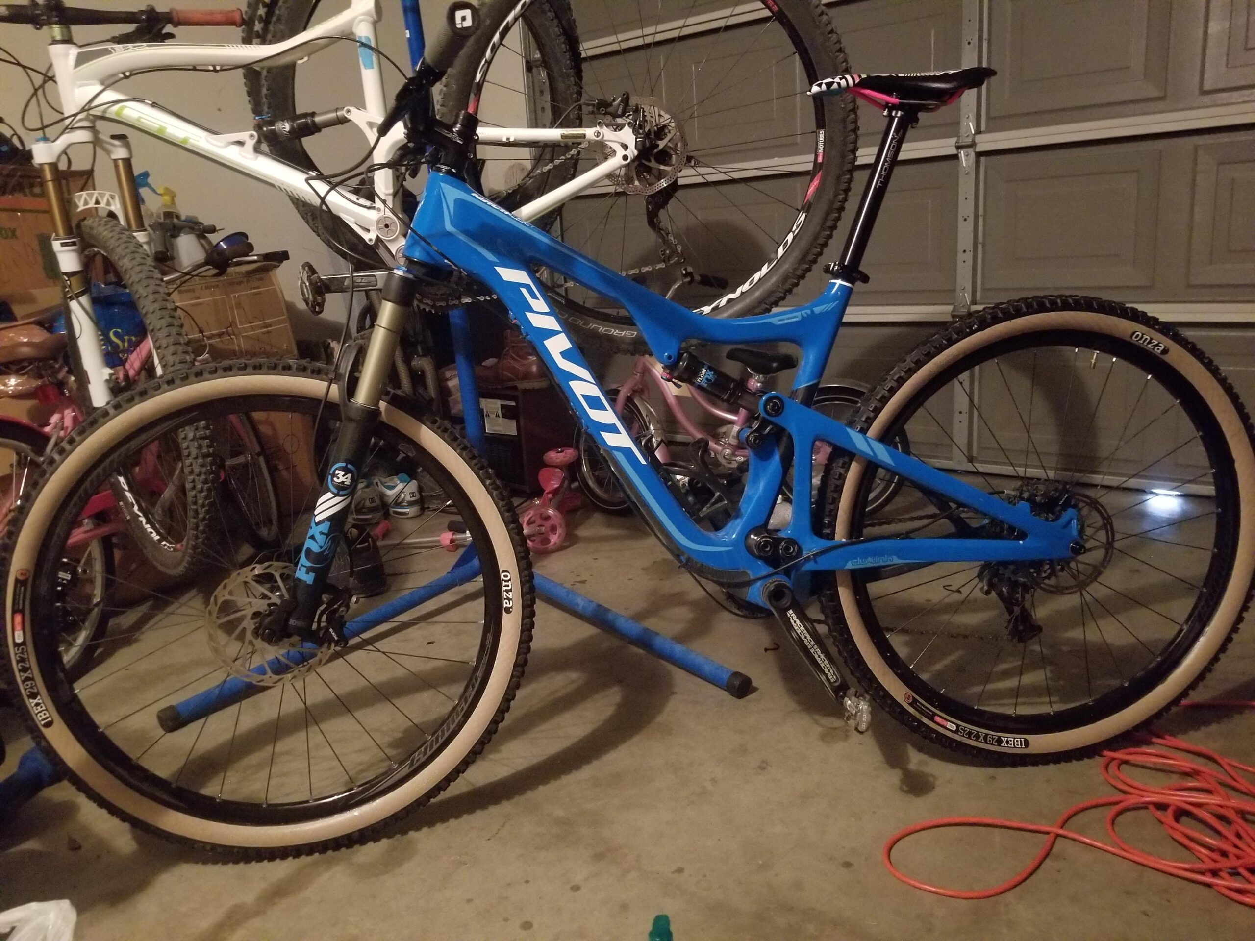 Pivot Trail 429: A blue mountain bike is displayed in a garage, positioned on a repair stand. The bike features a sleek design with a prominent "Pivot" logo, equipped with front suspension and knobby tires. In the background, a white bike frame can be seen hanging on the wall, along with various tools and equipment. The garage floor is cluttered with bike parts and accessories, indicating an active cycling space.