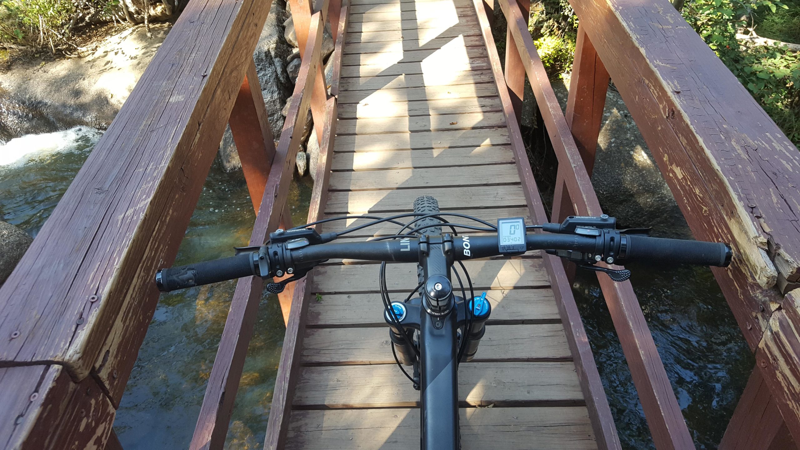A view from above a bicycle's handlebars, showing a wooden bridge over a clear stream. The bridge is rustic, with visible wear and natural surroundings of greenery and water. Curt Gowdy State Park mountain bike trail.
