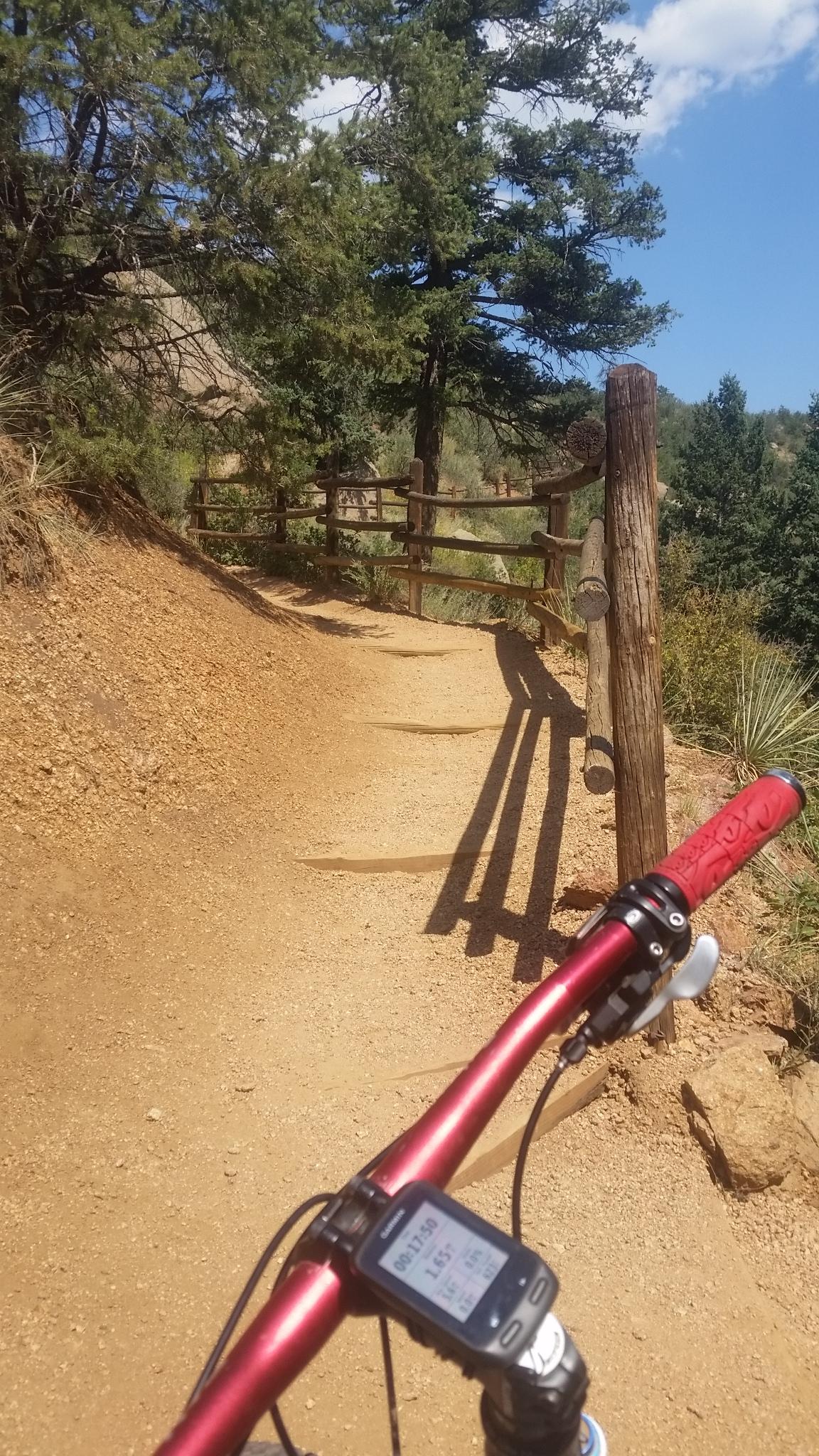 A close-up view of a mountain bike handlebar and digital display, overlooking a winding dirt trail surrounded by trees and wooden fencing. The scene is illuminated by bright sunlight, suggesting a clear day for outdoor biking. Barr Trail / Pikes Peak mountain bike trail.