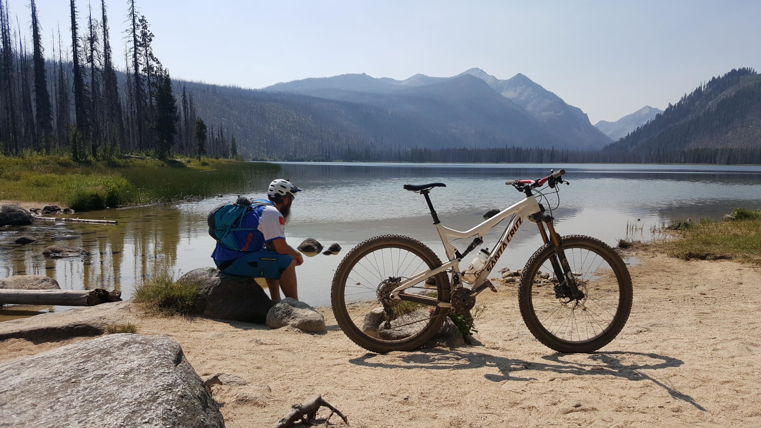 A cyclist sitting on a rock near a serene lake, reflecting on the water with mountains in the background. A white mountain bike is positioned in the foreground on sandy ground, surrounded by rocks and greenery. The scene captures a tranquil moment in a natural setting, with trees partially visible in the background. Loon Lake Trail mountain bike trail.