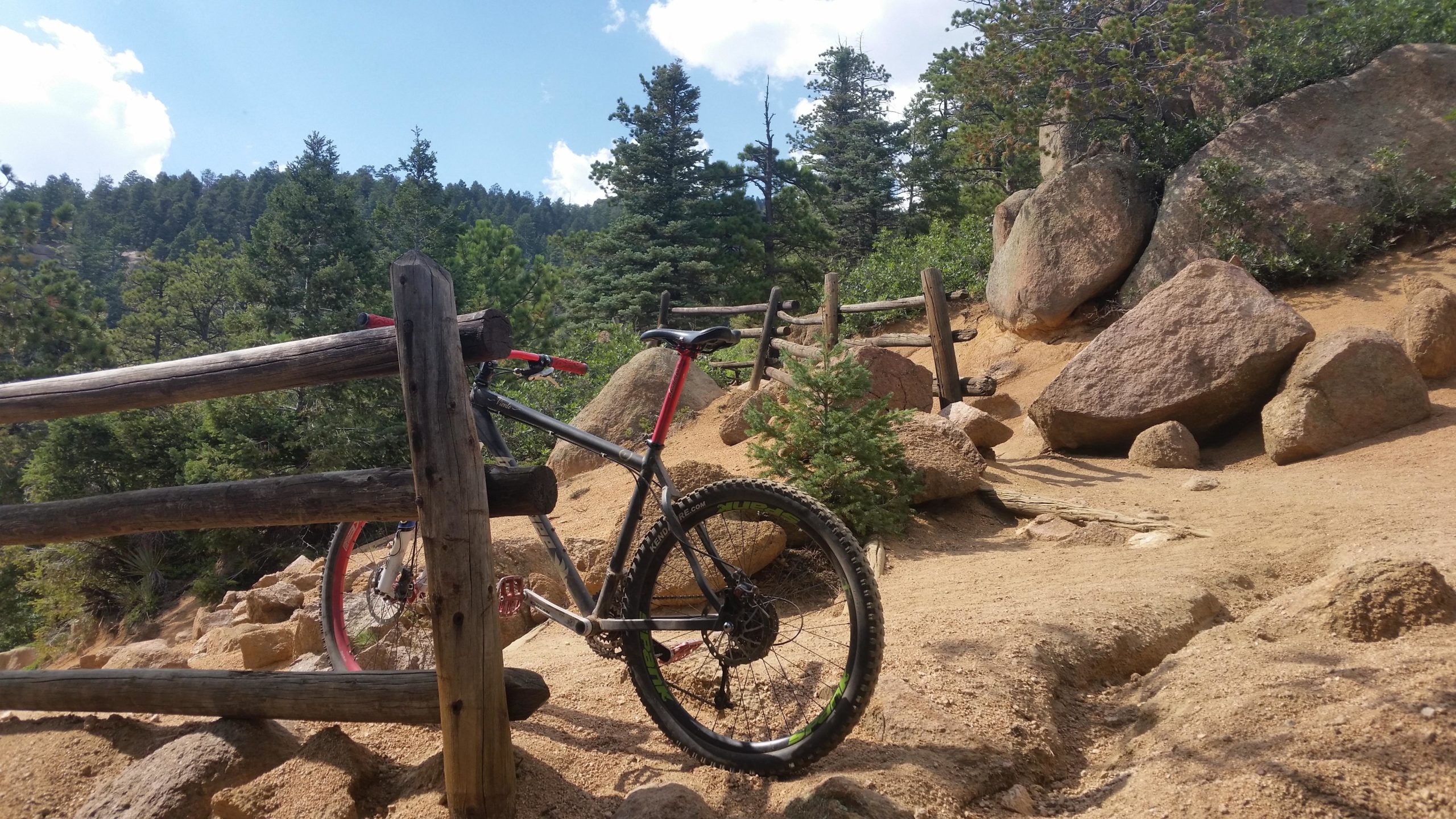 A mountain bike leans against a wooden fence along a rugged, sandy trail surrounded by trees and large rocks. The background features a pine forest under a partly cloudy sky. Barr Trail / Pikes Peak mountain bike trail.